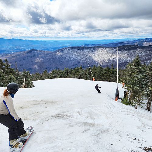 Two snowboarders glide down a slope with mountain views and partly sunny skies